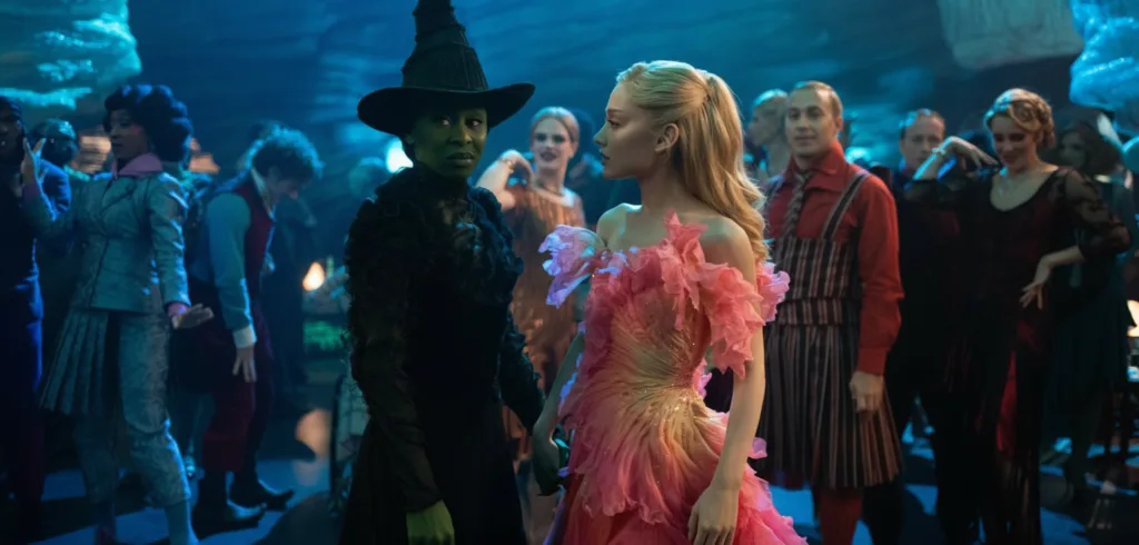 Crowded fantasy ballroom where a green-skinned woman in a black dress and a blonde woman in a pink ruffled gown stand together among dancing guests under blue lighting.”
